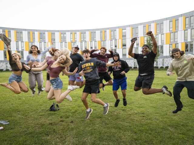 A group of Rowan University students jump and pose together on a grassy lawn outside a campus residence hall.