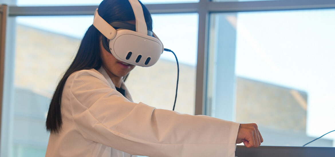 Rowan ECE major Gabriella P. concentrates on using a VR headset while moving her right arm. She is wearing a white lab coat.