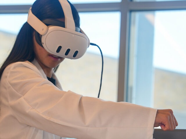 Rowan ECE major Gabriella P. concentrates on using a VR headset while moving her right arm. She is wearing a white lab coat.