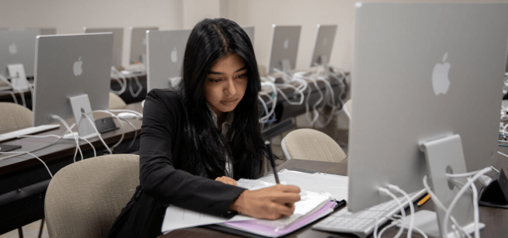 A student takes notes while working at a computer in a cybersecurity learning environment.