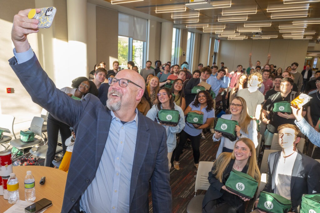 Business executive taking a selfie with a large group of Rowan students holding green V8 bags in a classroom.