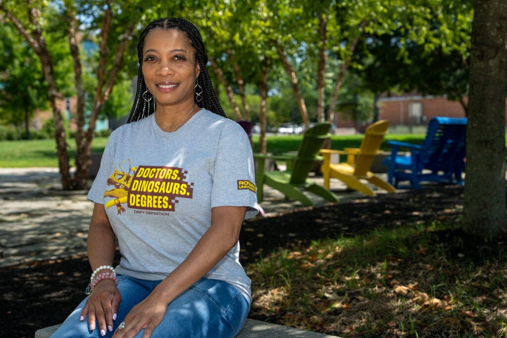 Dr. Clarke sits outdoors smiling at the camera while wearing a “Doctors, Dinosaurs, Degrees” T-shirt.