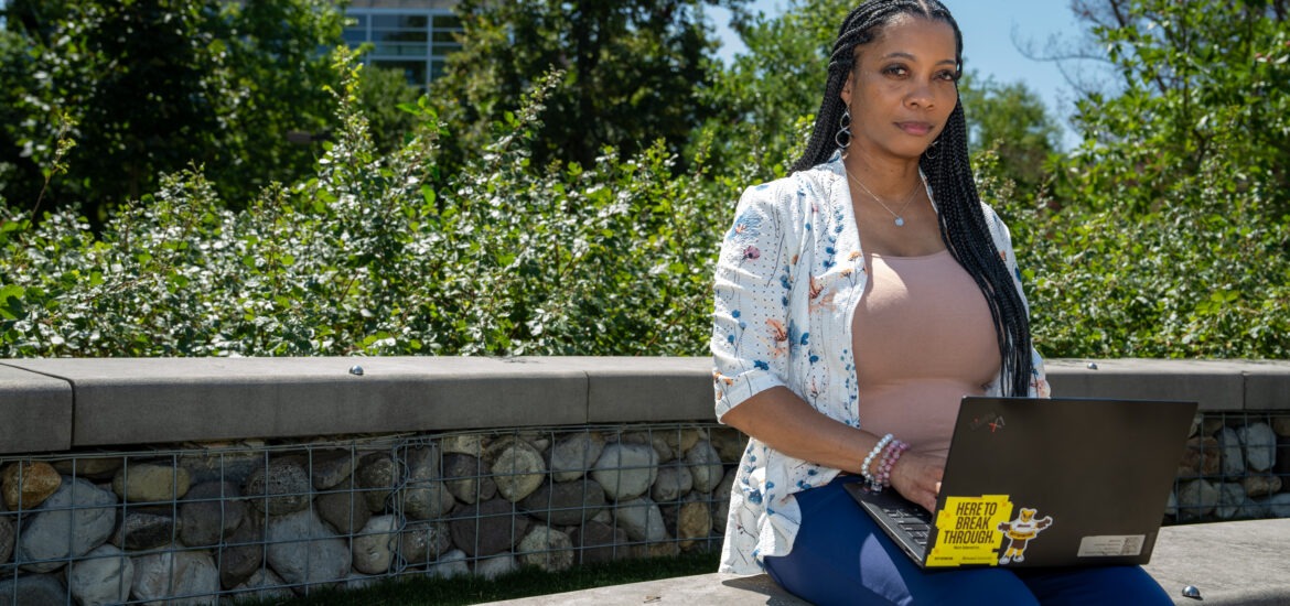 Jametta C. sits outdoors on a stone wall with an open laptop on her lap.