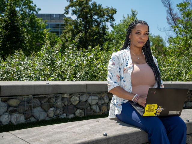 Jametta C. sits outdoors on a stone wall with an open laptop on her lap.