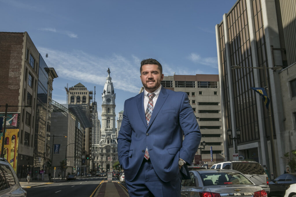 Man in a blue suit standing on a Philadelphia street with City Hall visible behind him.