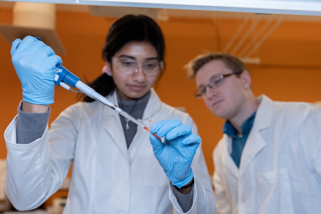Student wearing safety glasses and blue gloves using a pipette in a laboratory while another student observes.