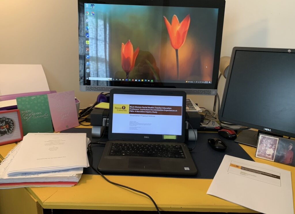 Jametta C.’s laptop displays her virtual dissertation defense, with an external monitor behind it and a framed photo of her parents beside the computer.