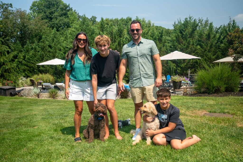 A family-provided photo, Rex sits in his grassy backyard with his human mom and dad, two human brothers and his fur brother Rocco.