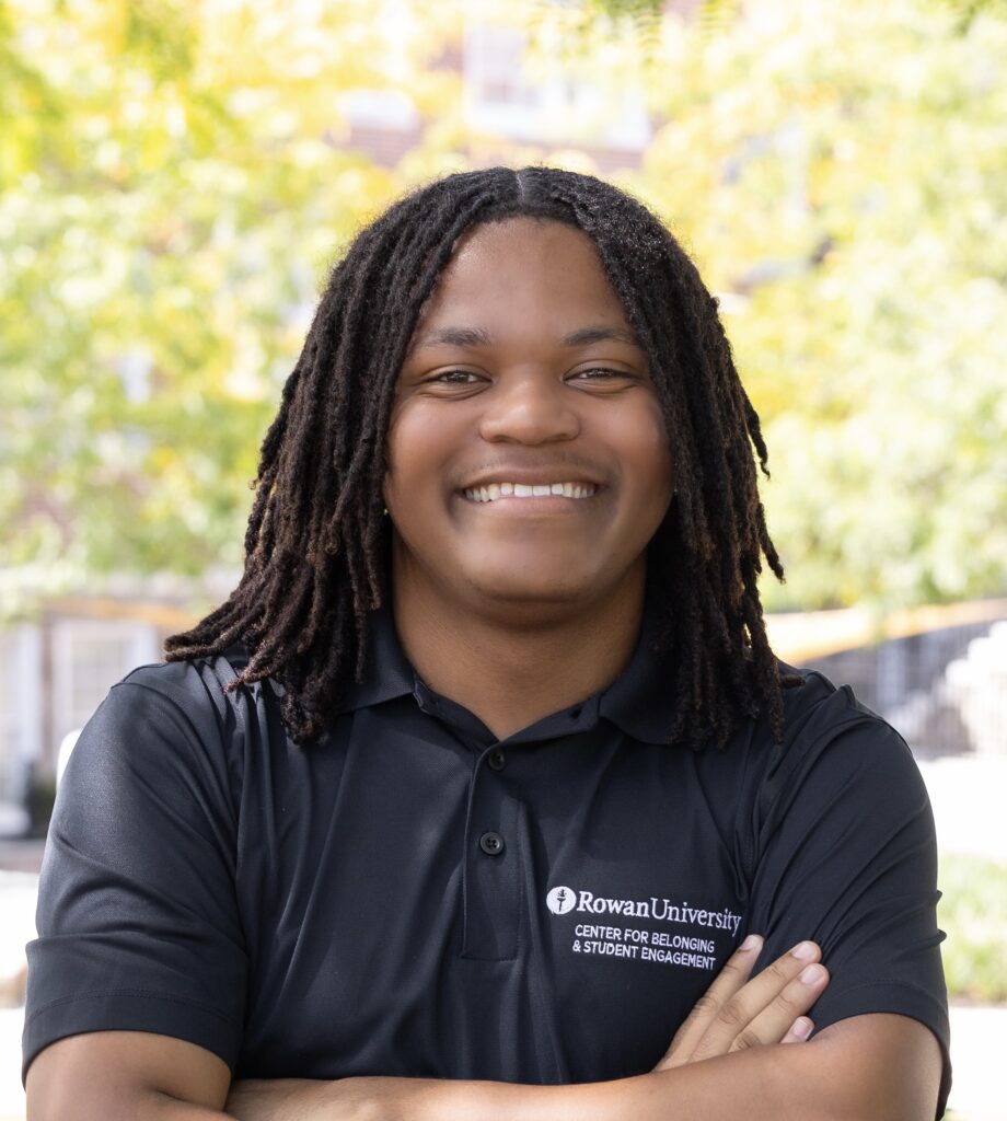 Student smiles with arms crossed outdoors while wearing a black Rowan University Center for Belonging & Student Engagement polo shirt.
