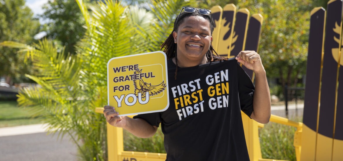 Student smiles outdoors while holding a “We’re Grateful for You” sign and wearing a black T-shirt that reads “First Gen” repeatedly.