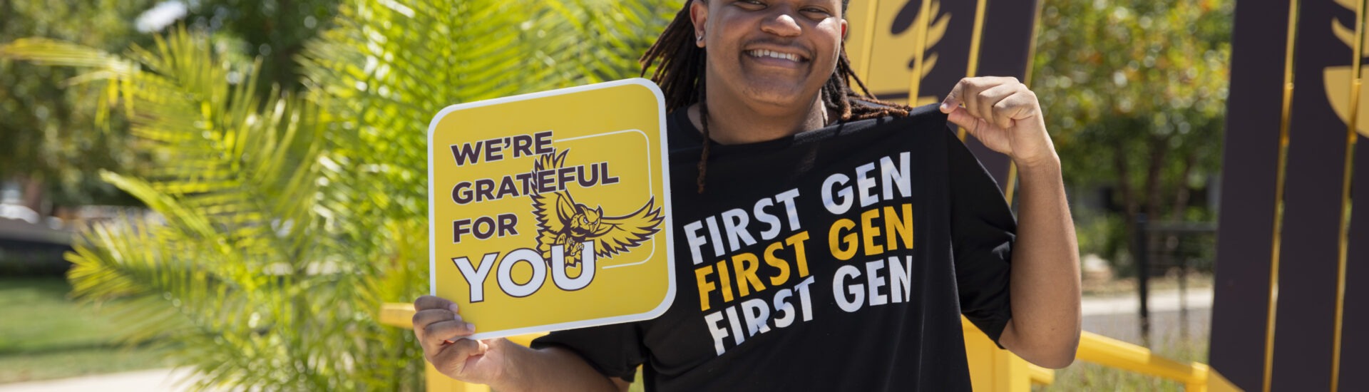 Student smiles outdoors while holding a “We’re Grateful for You” sign and wearing a black T-shirt that reads “First Gen” repeatedly.