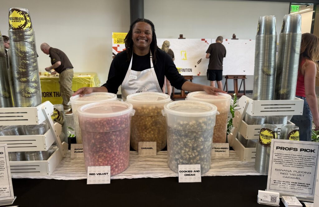Student stands behind The Ballard Bakery table at Defy Definition LIVE event with several large containers of colorful gourmet popcorn displayed for customers.